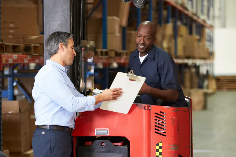 Two colleagues reviewing a clipboard beside a forklift in a warehouse